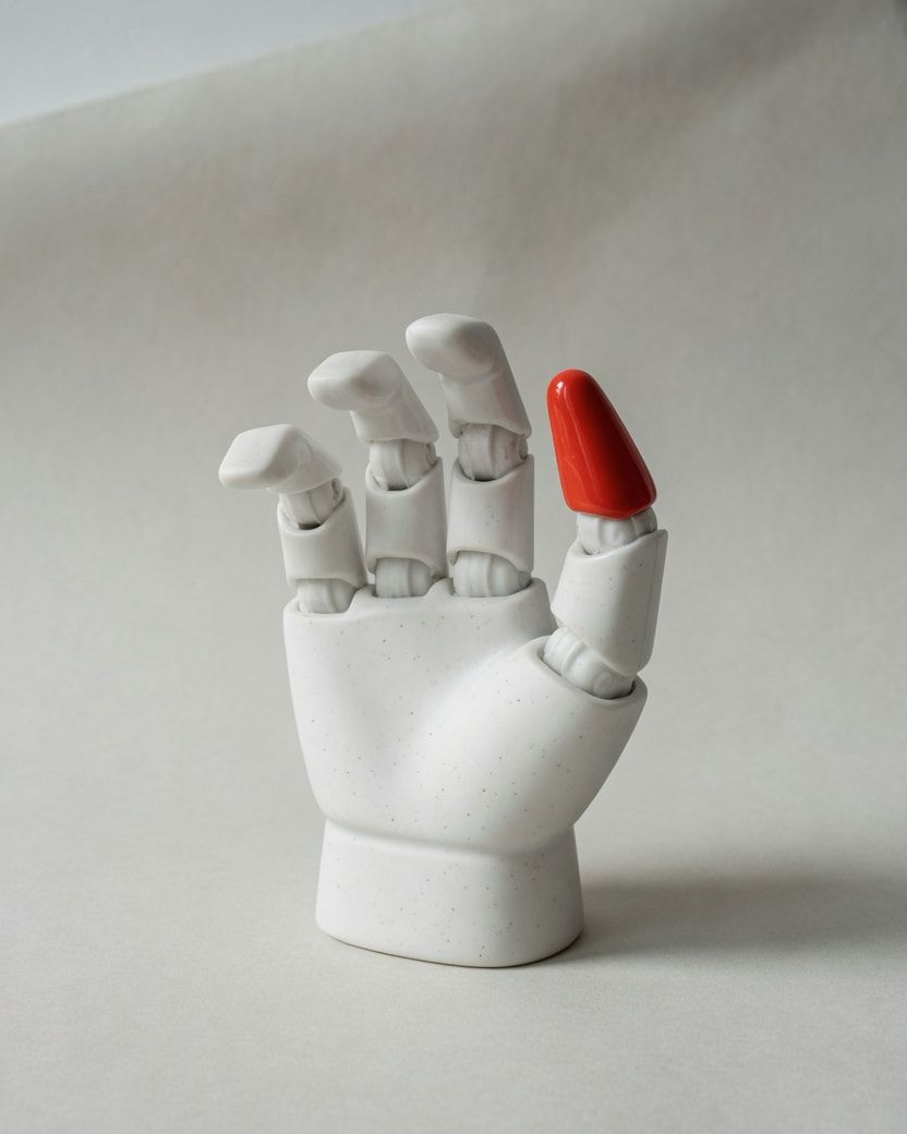 Studio photograph of a white porcelain robotic gripper with one vermilion fingertip.