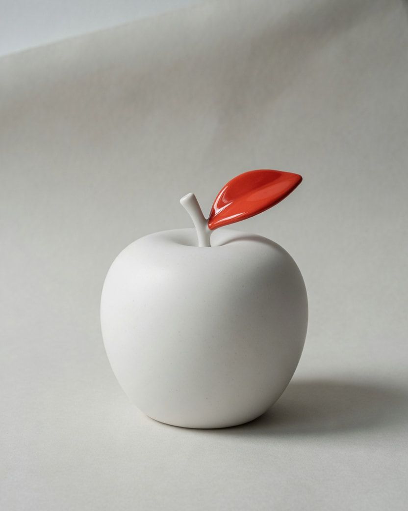 Studio photograph of a white porcelain apple with a vermilion leaf.