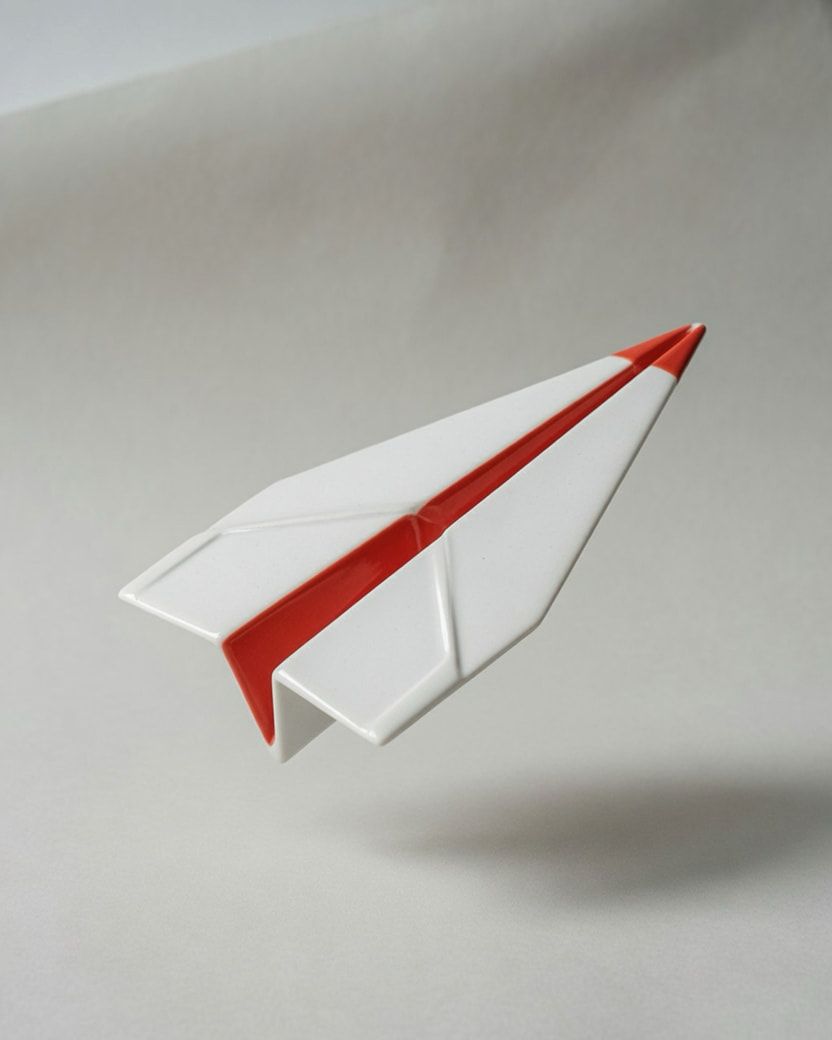 Studio photograph of a white porcelain paper airplane with a vermilion fold line.